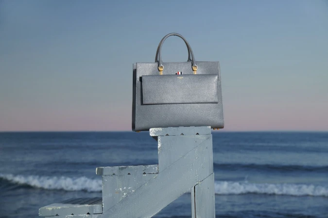 Grey leather tote bag with top handle and front flap pocket sitting on top of blue painted lifeguard stand with ocean and sky background.