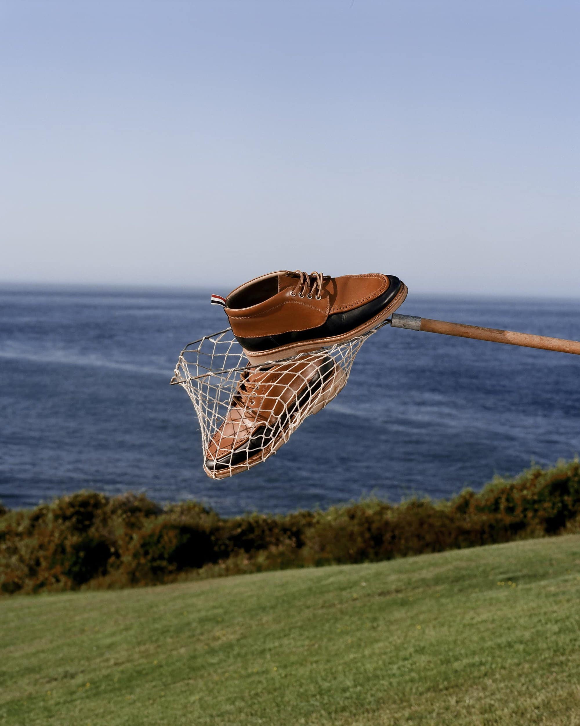 Brown leather boot held in fishing net on a wooden pole with ocean and green hillside background on a clear day, product photography.