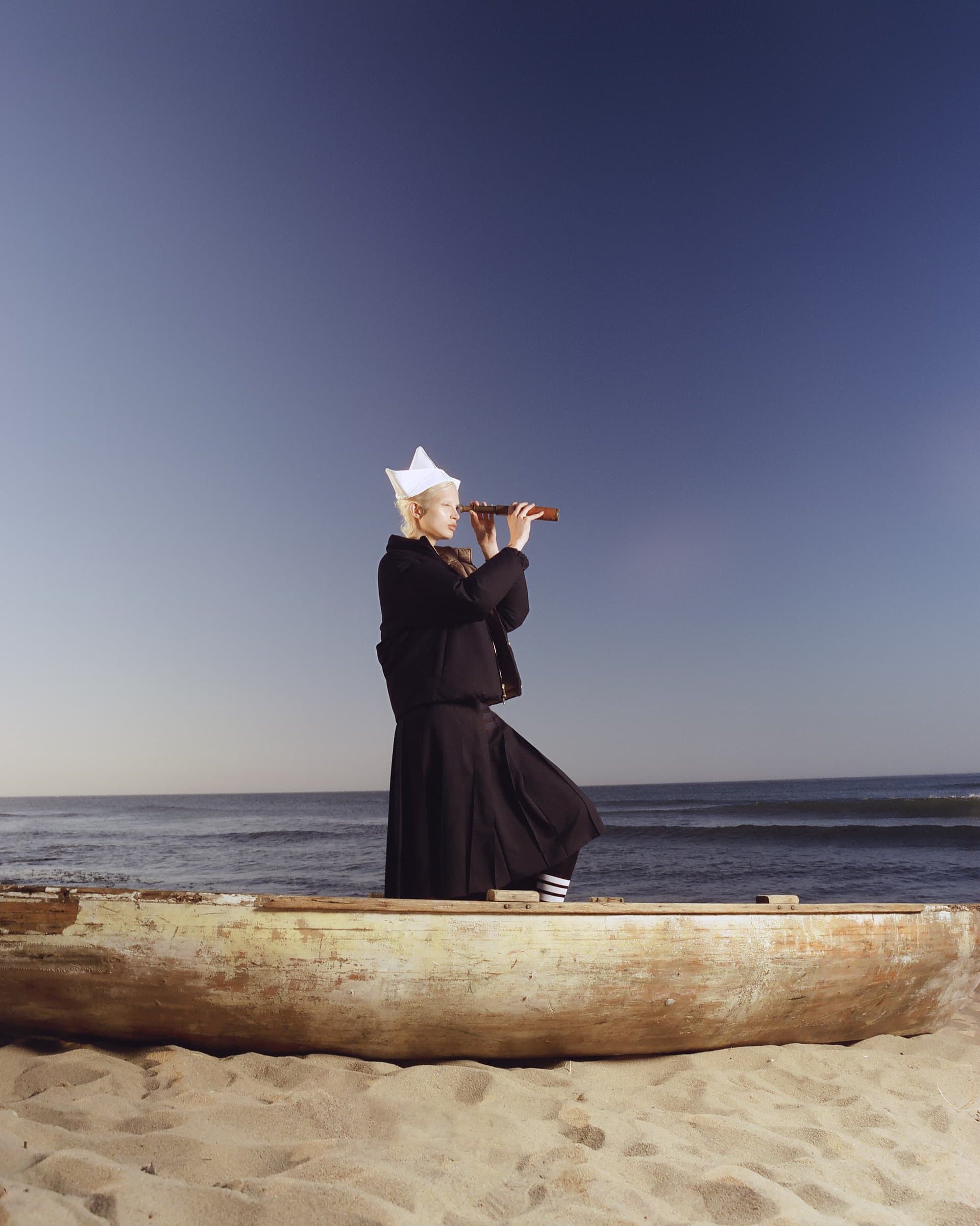Adult female standing in a weathered boat on a beach, wearing a white paper hat and black clothes, looking through a telescope.