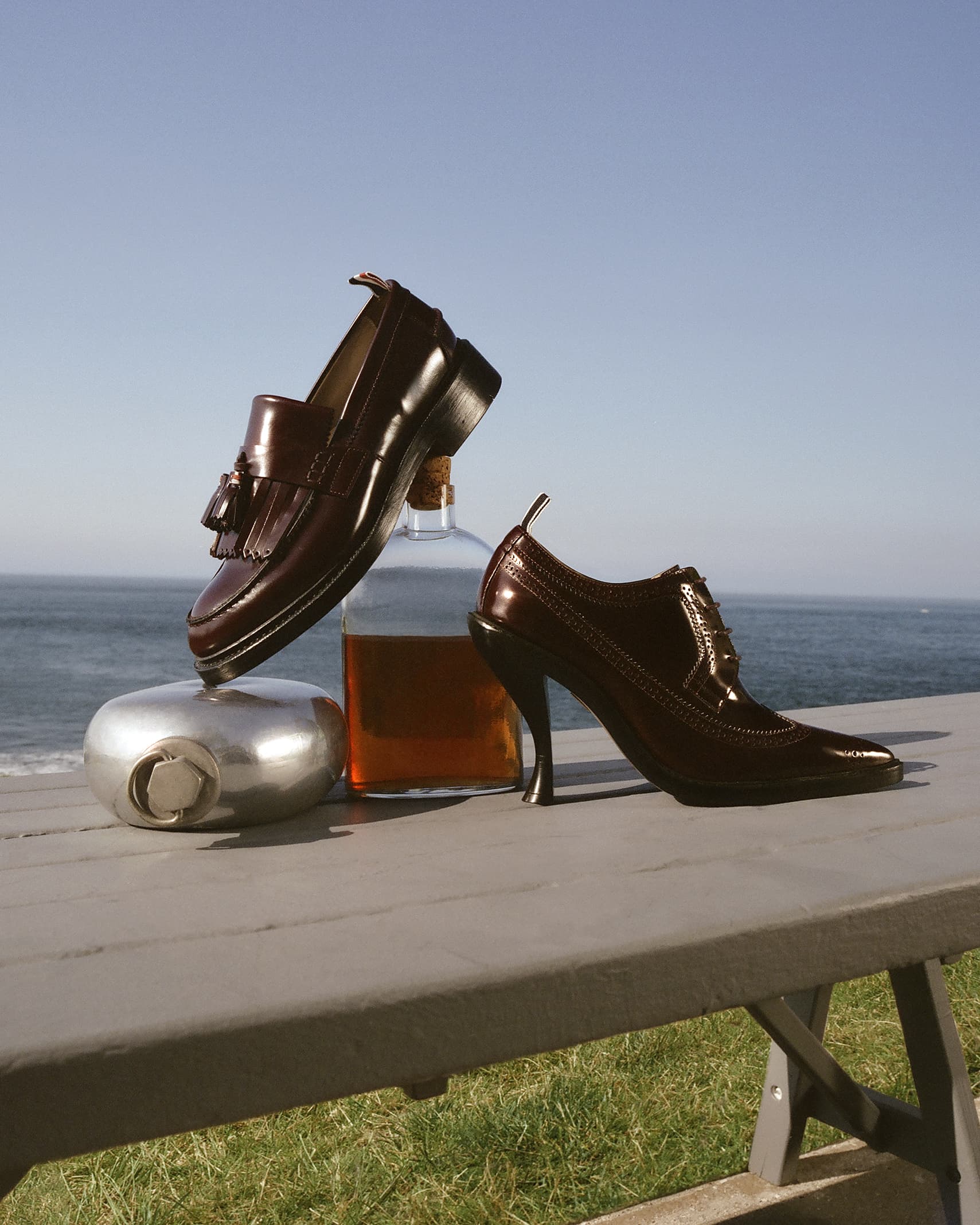 Still life shot of a loafer, heeled shoe, bottle of liquor, and silver flask on a gray surface with a coastal view backdrop.