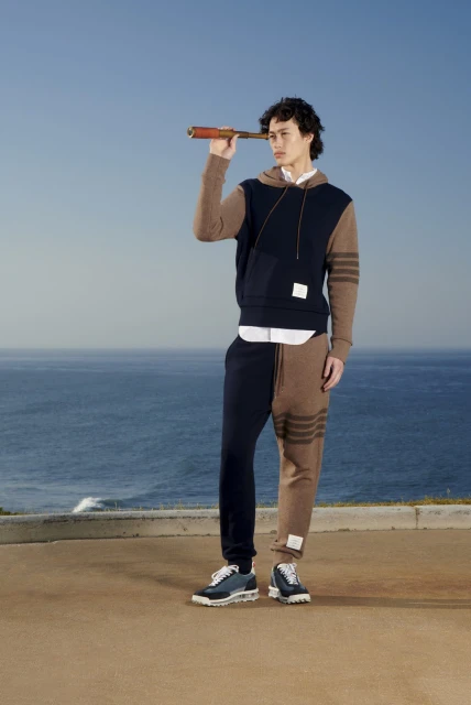 Man in color-blocked hoodie and sweatpants holds telescope on a coastal overlook with blue sky and ocean backdrop. Fashion shot.