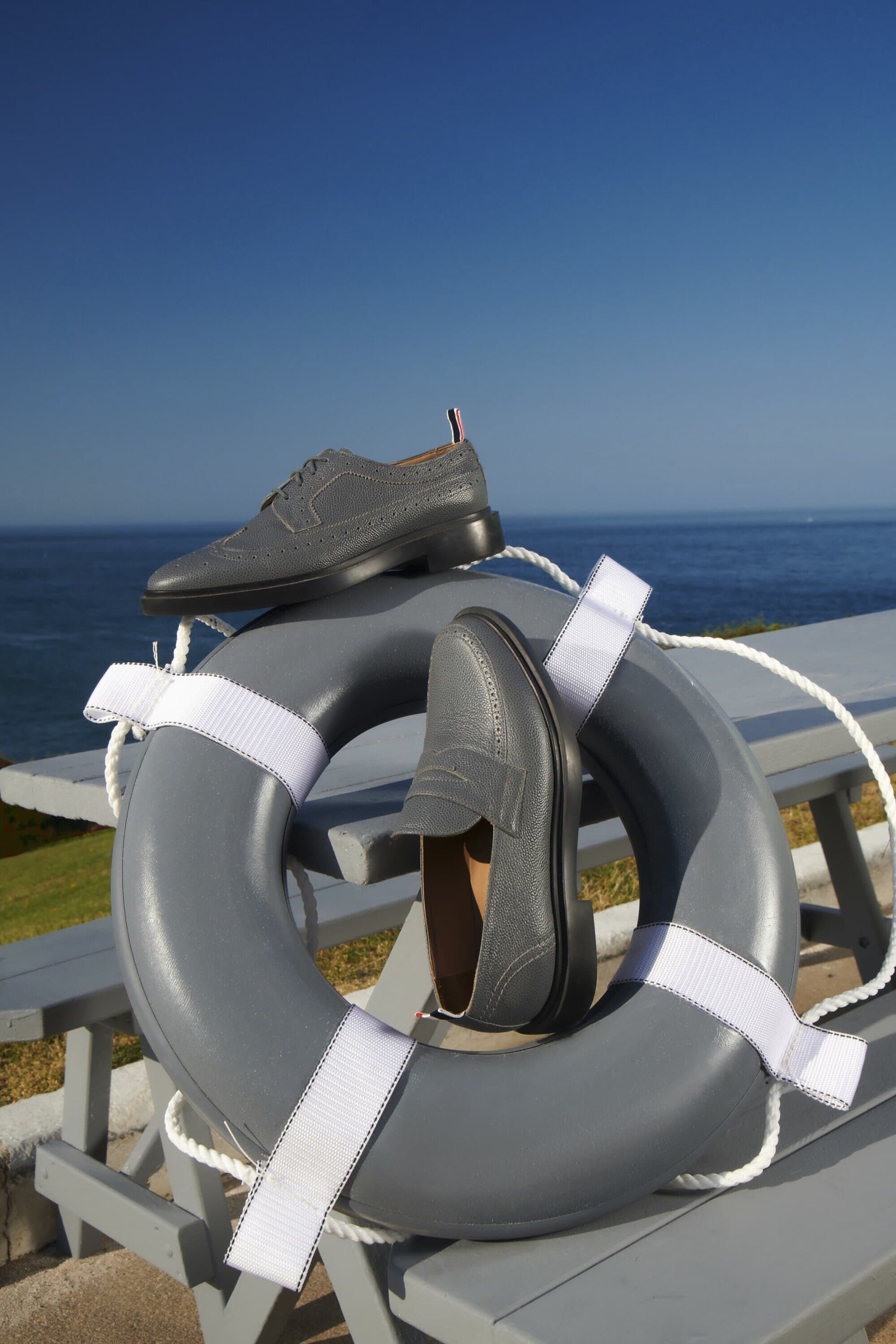Grey leather loafers and brogues sit on a life preserver on a grey wooden bench with a blue ocean and sky backdrop.