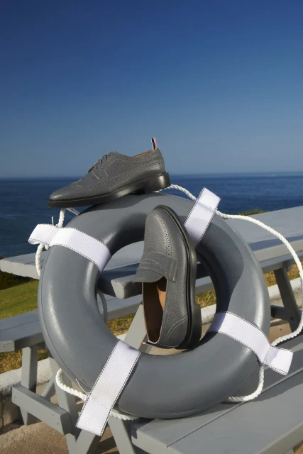 Grey leather loafers and brogues sit on a life preserver on a grey wooden bench with a blue ocean and sky backdrop.