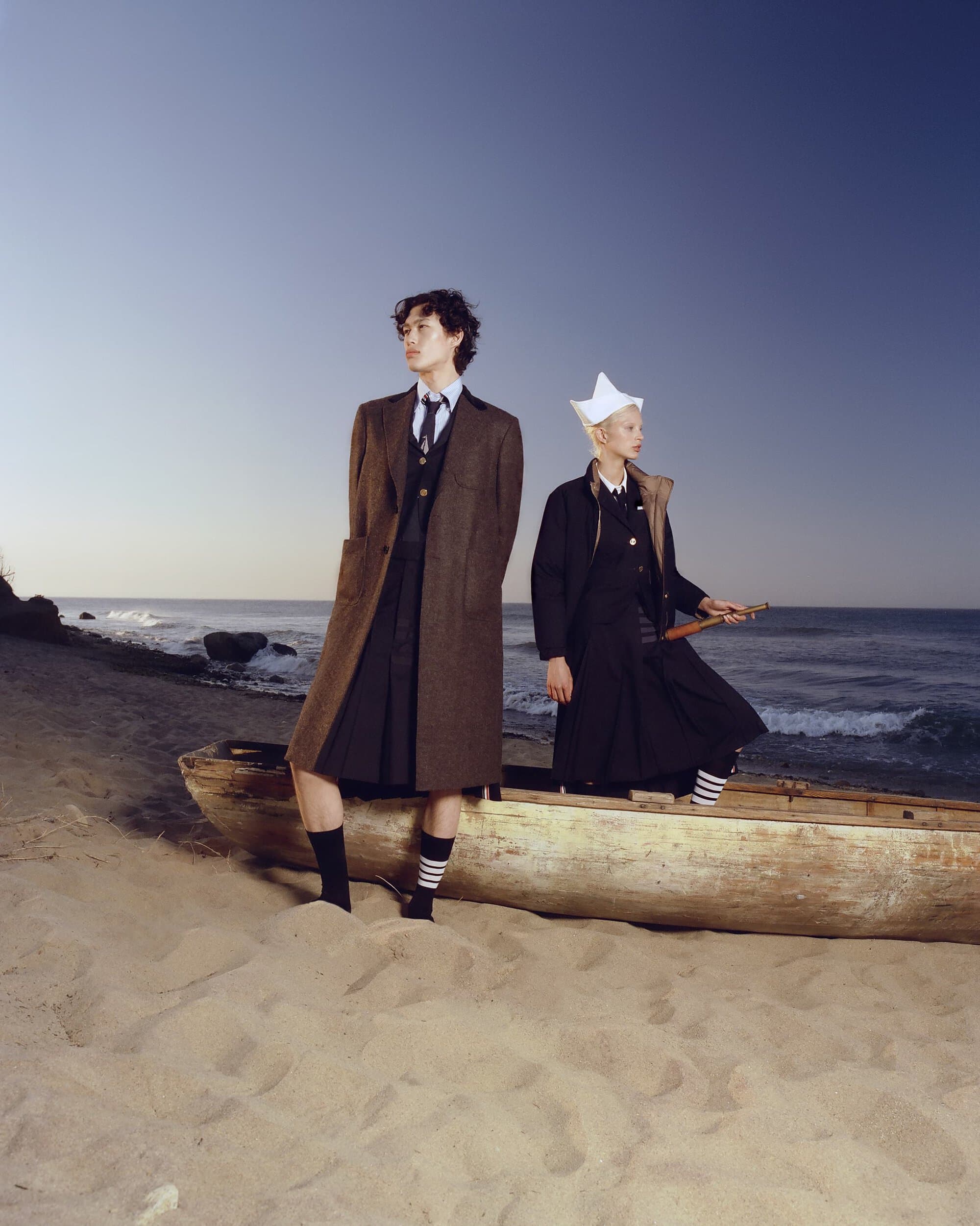 Two people in fashion-forward formalwear and paper hat stand next to a boat on a sandy beach with calm ocean and a clear blue sky.