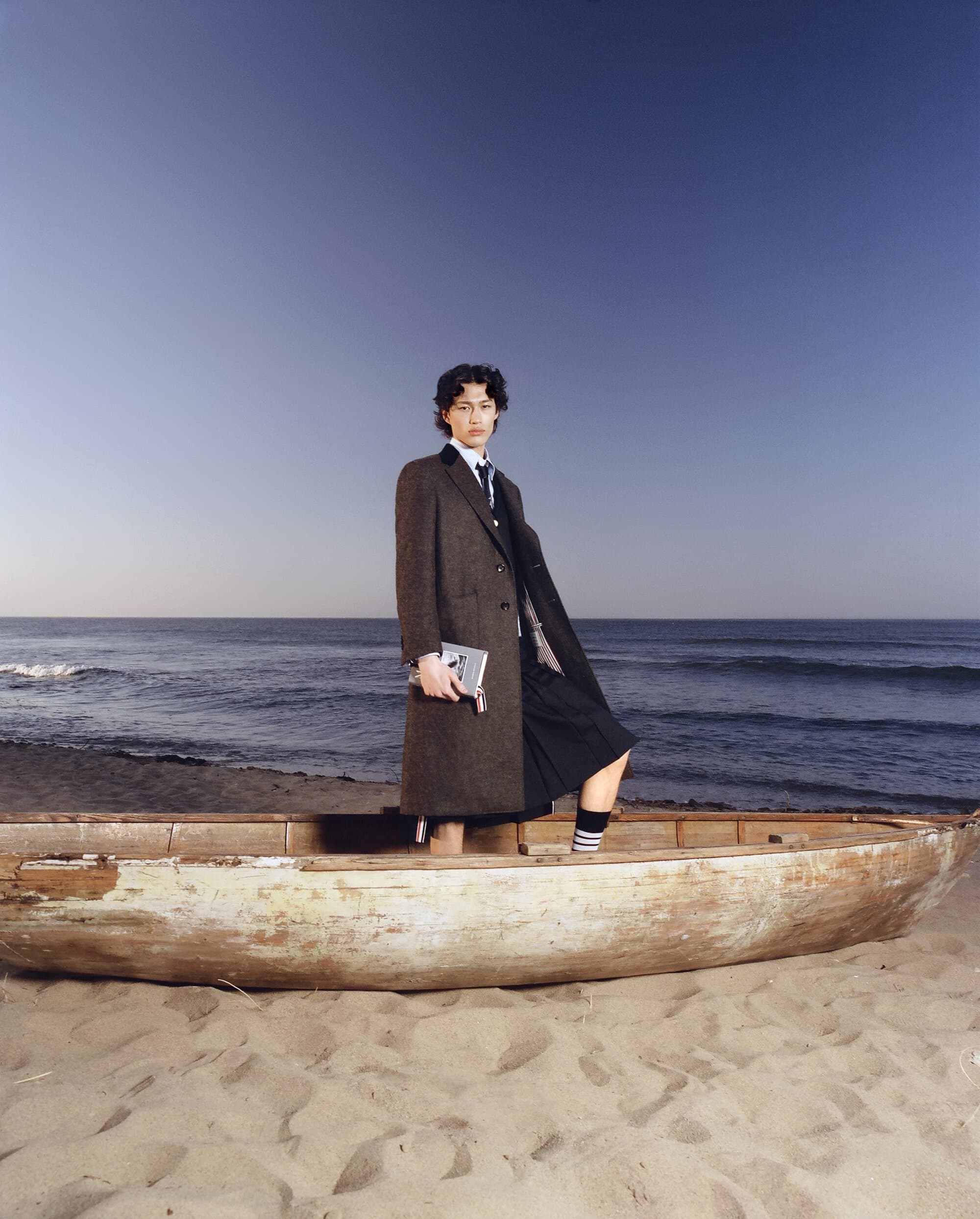 Man in a tweed coat and kilt stands in a weathered rowboat on a sandy beach with the ocean and blue sky visible in the background.