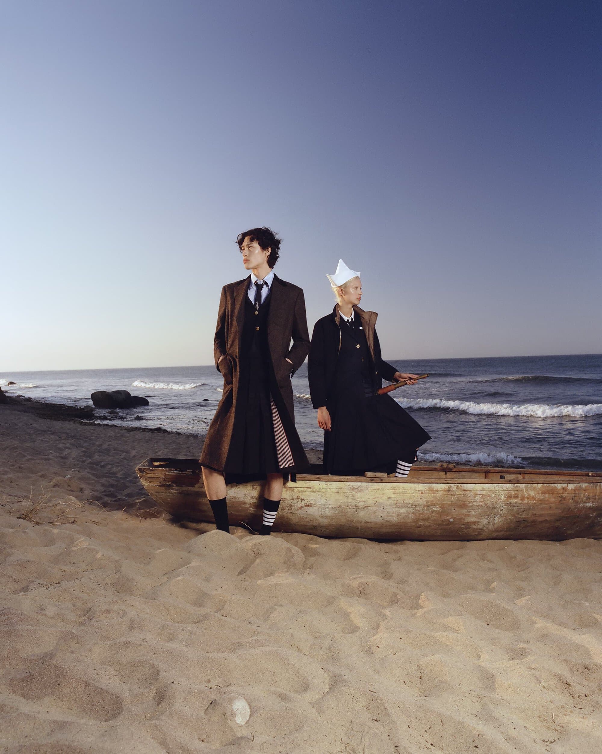 Two people stand in a boat on a sandy beach next to the ocean. The person to the left wears a kilt and the other a white hat.