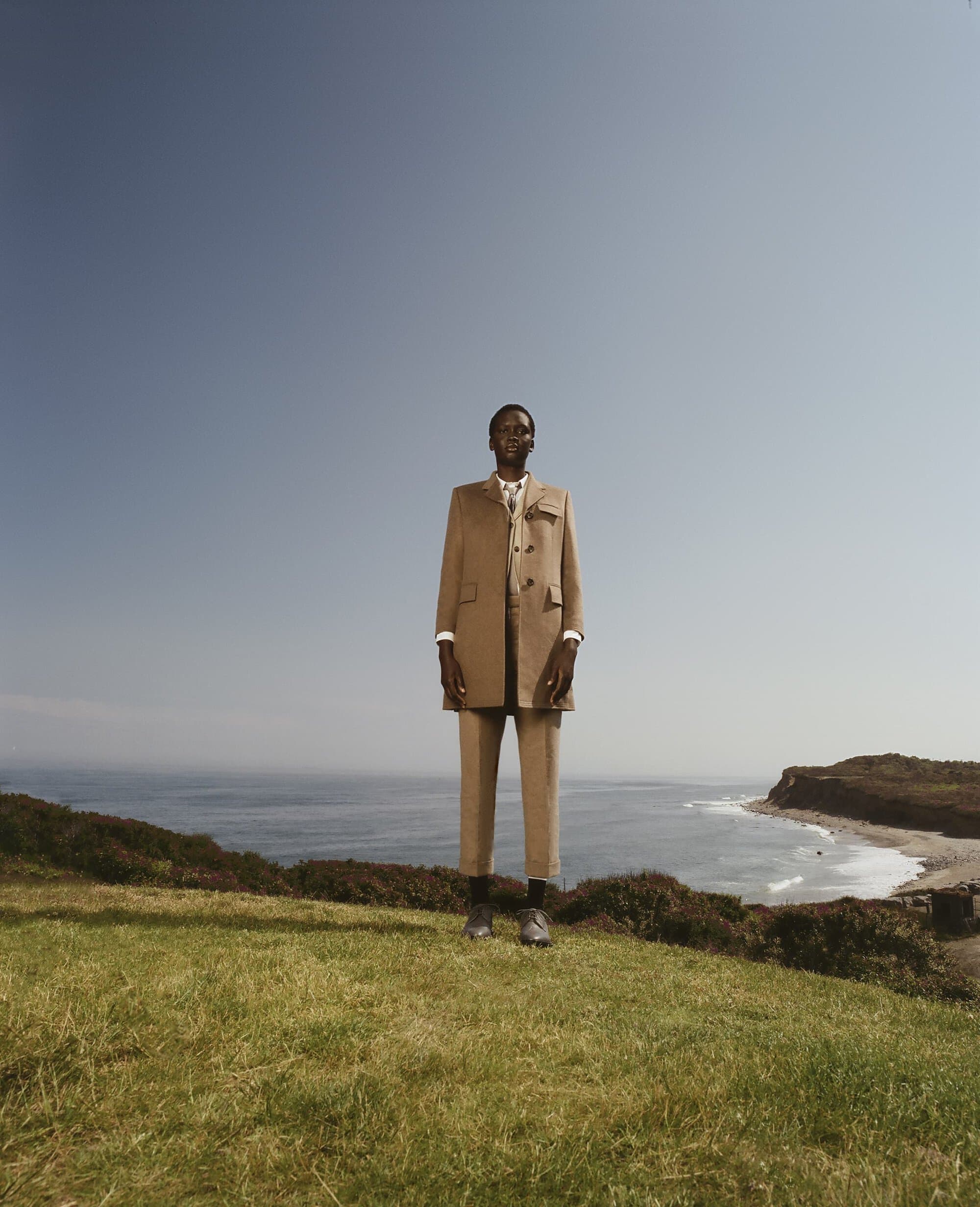 An adult male model poses in an outdoor fashion editorial on a grassy hill overlooking the ocean, wearing a tan suit and dress shoes.