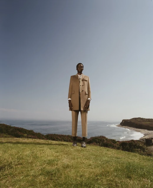 An adult male model poses in an outdoor fashion editorial on a grassy hill overlooking the ocean, wearing a tan suit and dress shoes.