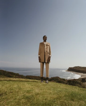 An adult male model poses in an outdoor fashion editorial on a grassy hill overlooking the ocean, wearing a tan suit and dress shoes.