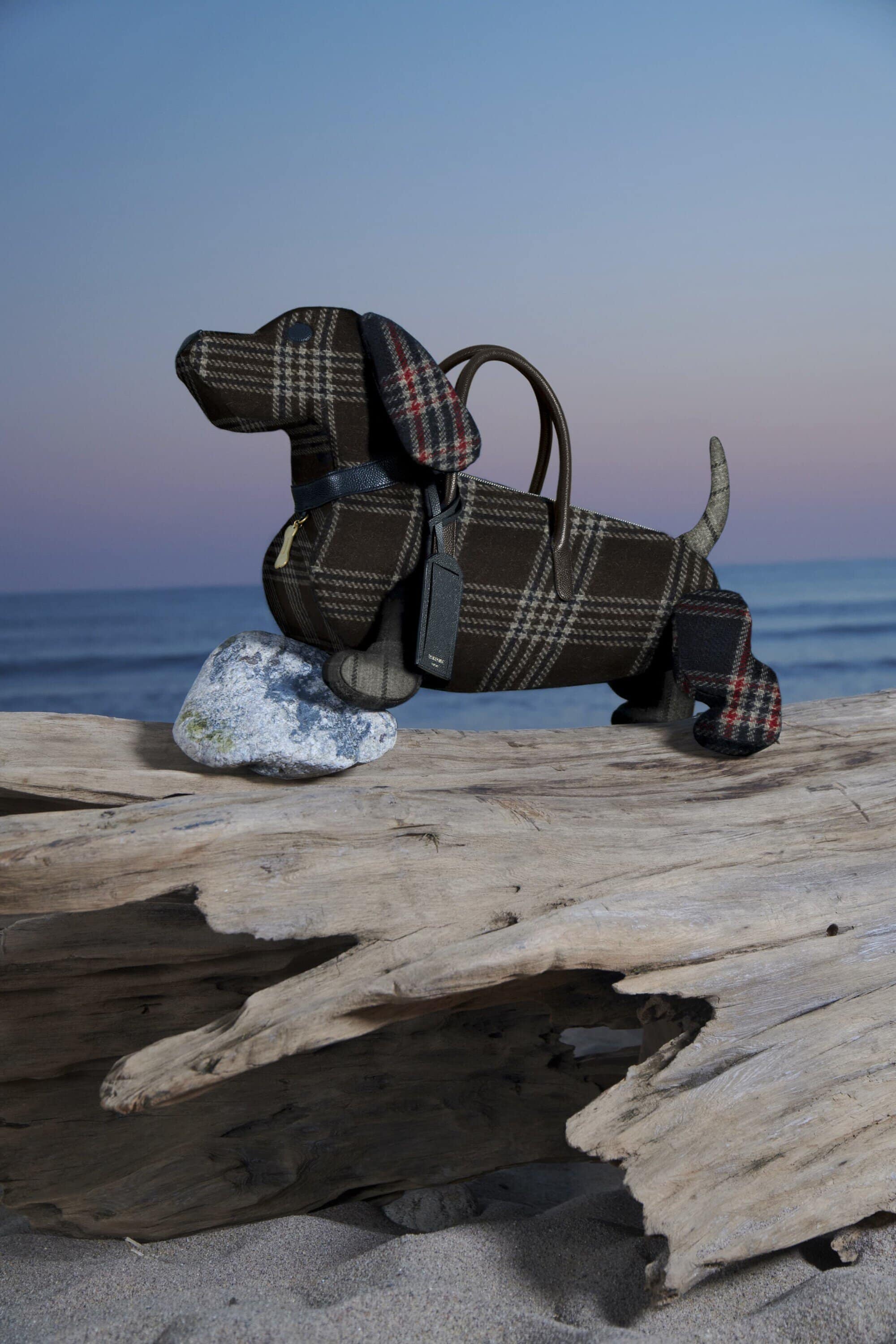 Dog-shaped plaid handbag on a weathered log, set against a serene beach backdrop with a soft blue sky.