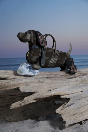 Dog-shaped plaid handbag on a weathered log, set against a serene beach backdrop with a soft blue sky.