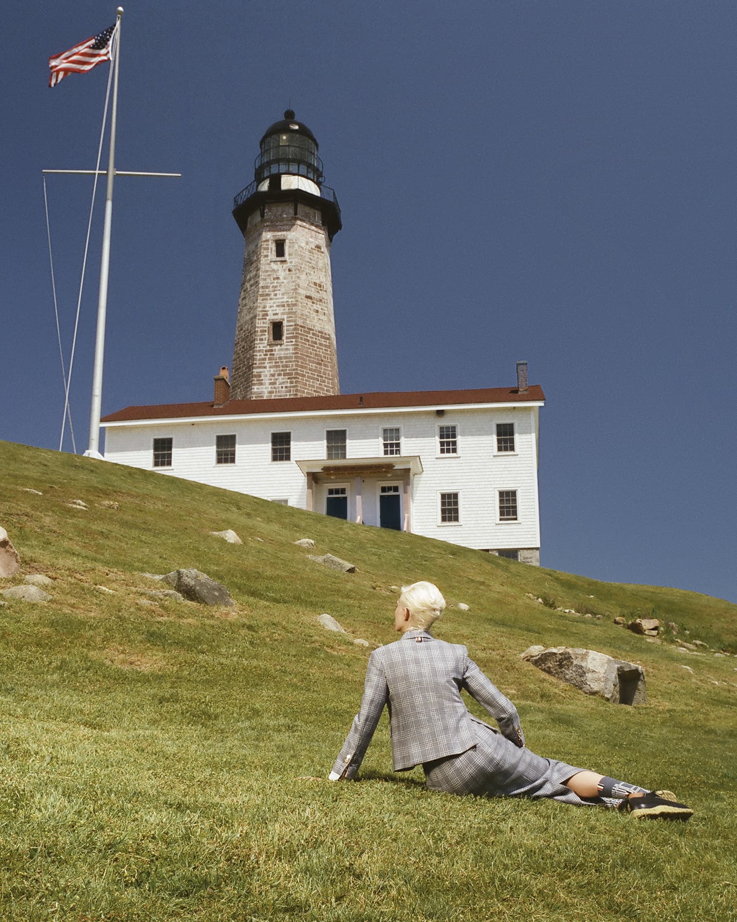 Person in suit lounges on grassy hill in front of white lighthouse under blue sky with USA flag; daytime, outdoor, landscape orientation.
