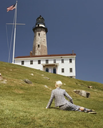 Person in suit lounges on grassy hill in front of white lighthouse under blue sky with USA flag; daytime, outdoor, landscape orientation.