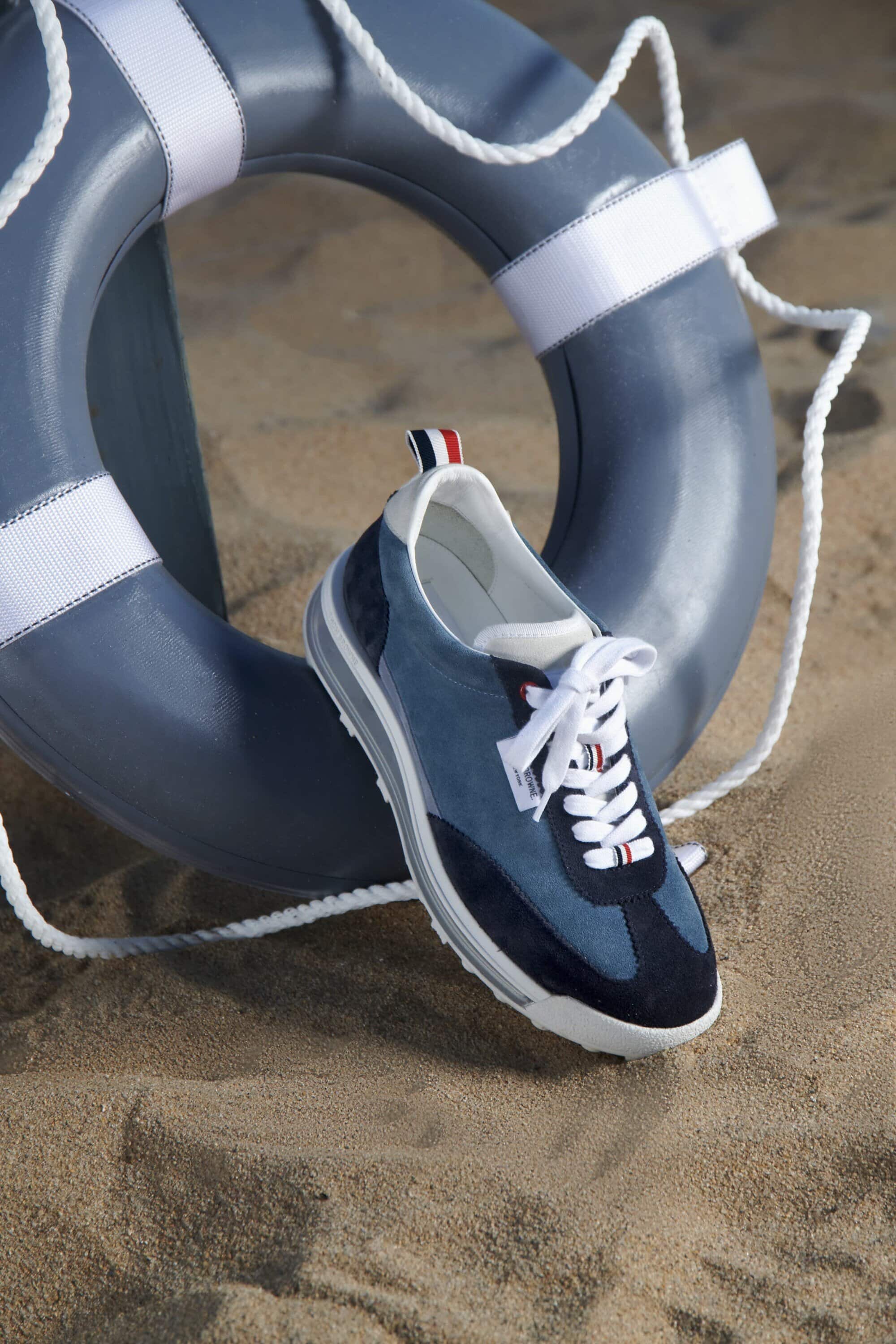 Blue casual sneaker on a sandy beach with a matching blue lifesaver ring in the background creates a summery footwear ad image.