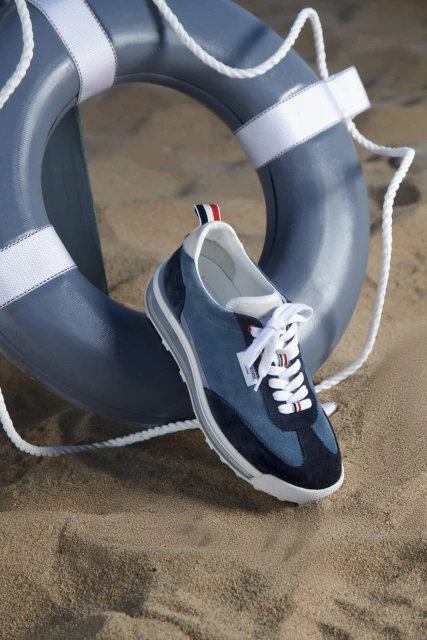 Blue casual sneaker on a sandy beach with a matching blue lifesaver ring in the background creates a summery footwear ad image.