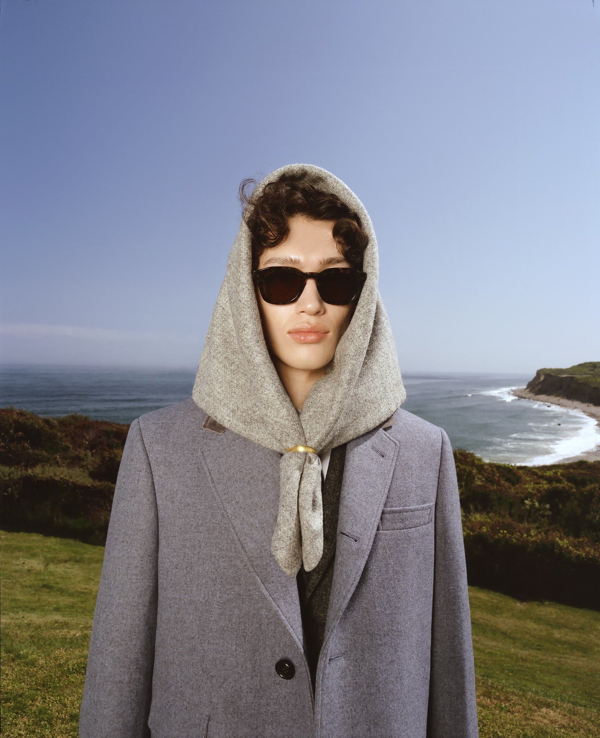 Person in sunglasses wearing a headscarf and coat, standing in front of a coastal landscape with a blue sky and ocean.