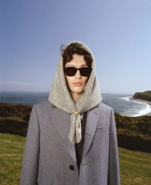 Person in sunglasses wearing a headscarf and coat, standing in front of a coastal landscape with a blue sky and ocean.