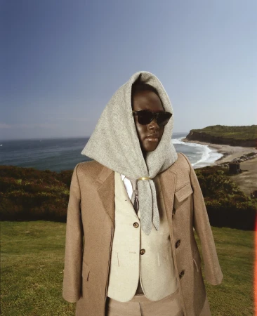A person wearing a grey wool headscarf and tan blazer over a beige vest, standing outdoors with a coastal landscape background.