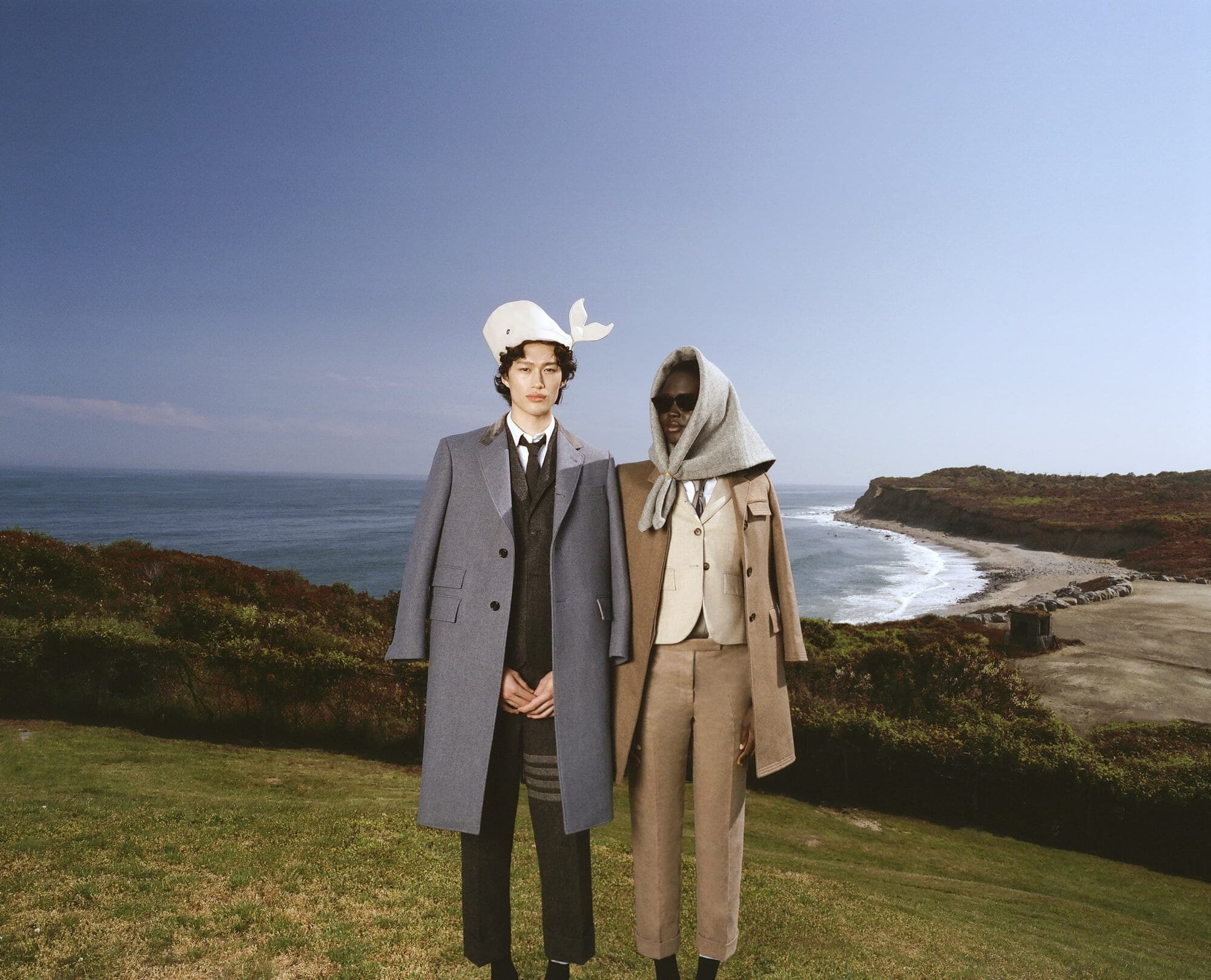 Two fashion models in stylish suits and hats stand on a grassy hill overlooking the ocean, blending fashion with scenic nature.