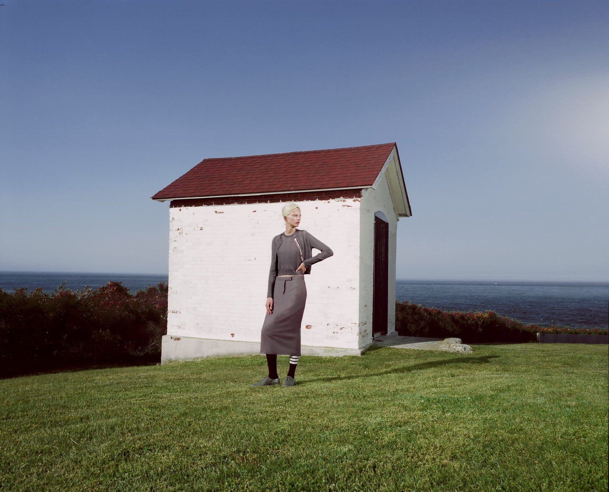A fair-skinned woman in a grey skirt and top stands on green grass near a white brick structure with a red roof, under a blue sky.