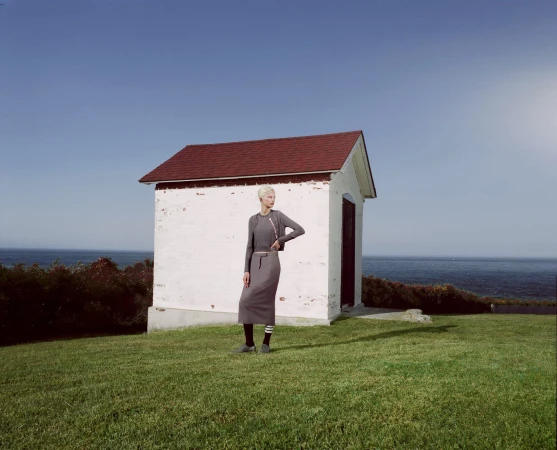 A fair-skinned woman in a grey skirt and top stands on green grass near a white brick structure with a red roof, under a blue sky.