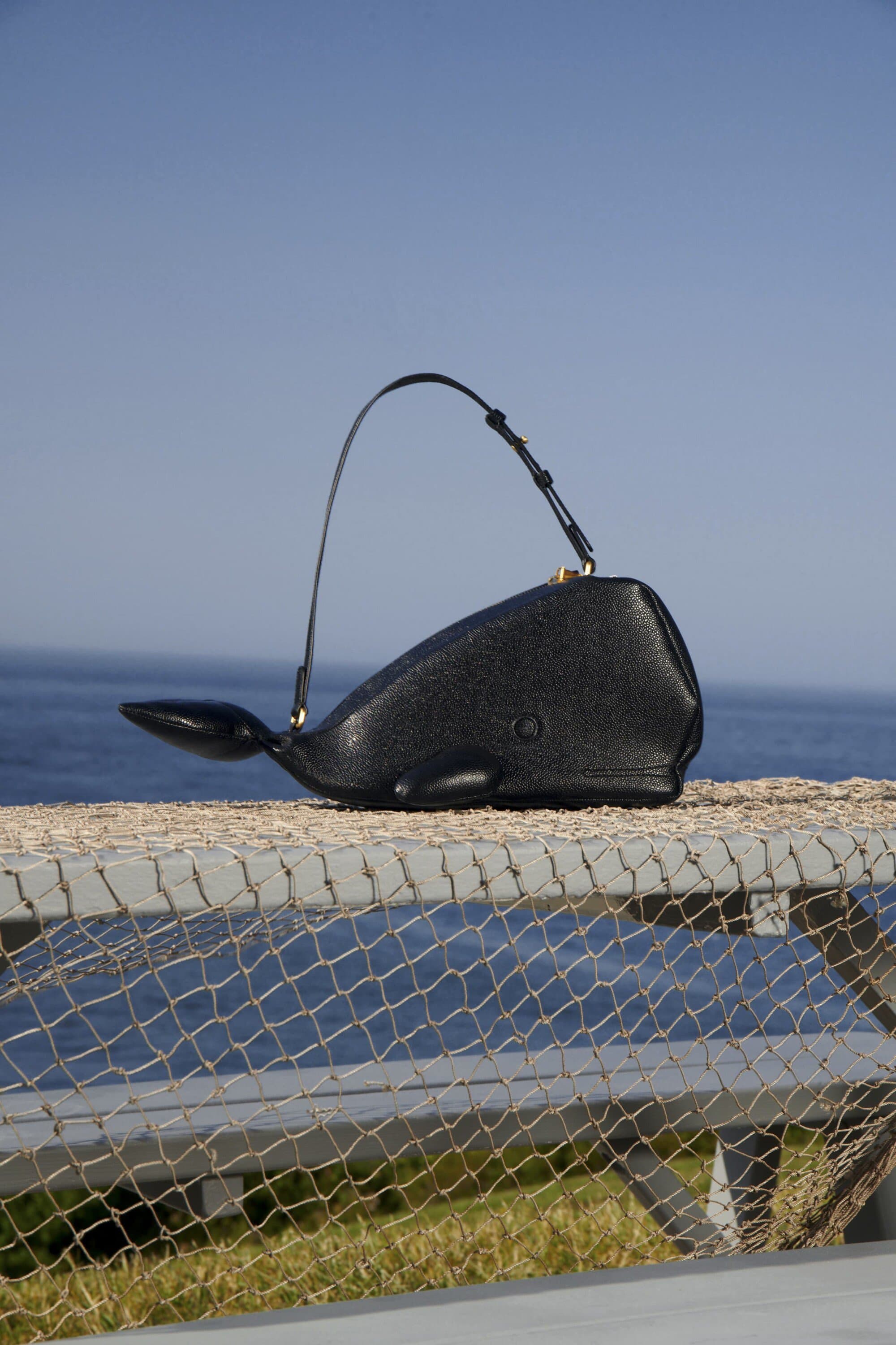 Black whale-shaped handbag with a curved handle sits on a metal railing with the ocean and blue sky in the background on a sunny day.