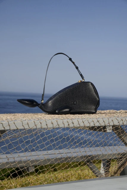 Black whale-shaped handbag with a curved handle sits on a metal railing with the ocean and blue sky in the background on a sunny day.