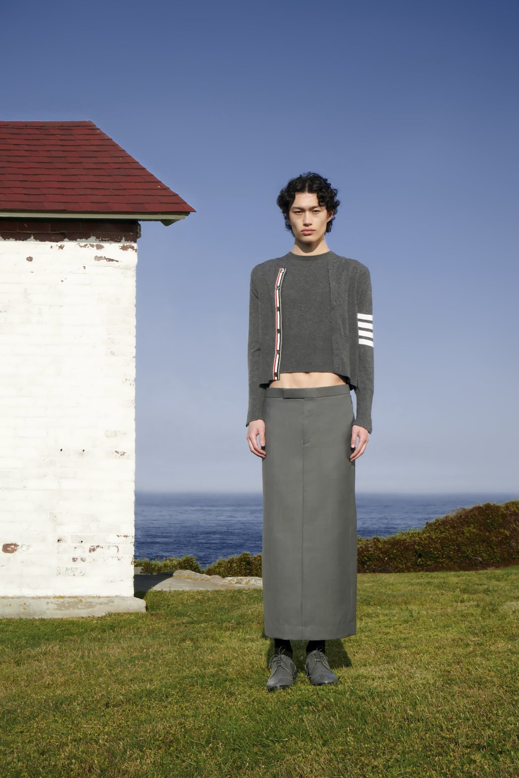 Young adult male poses in a grey knit sweater and long skirt on a lawn next to a weathered building and the ocean under a blue sky.