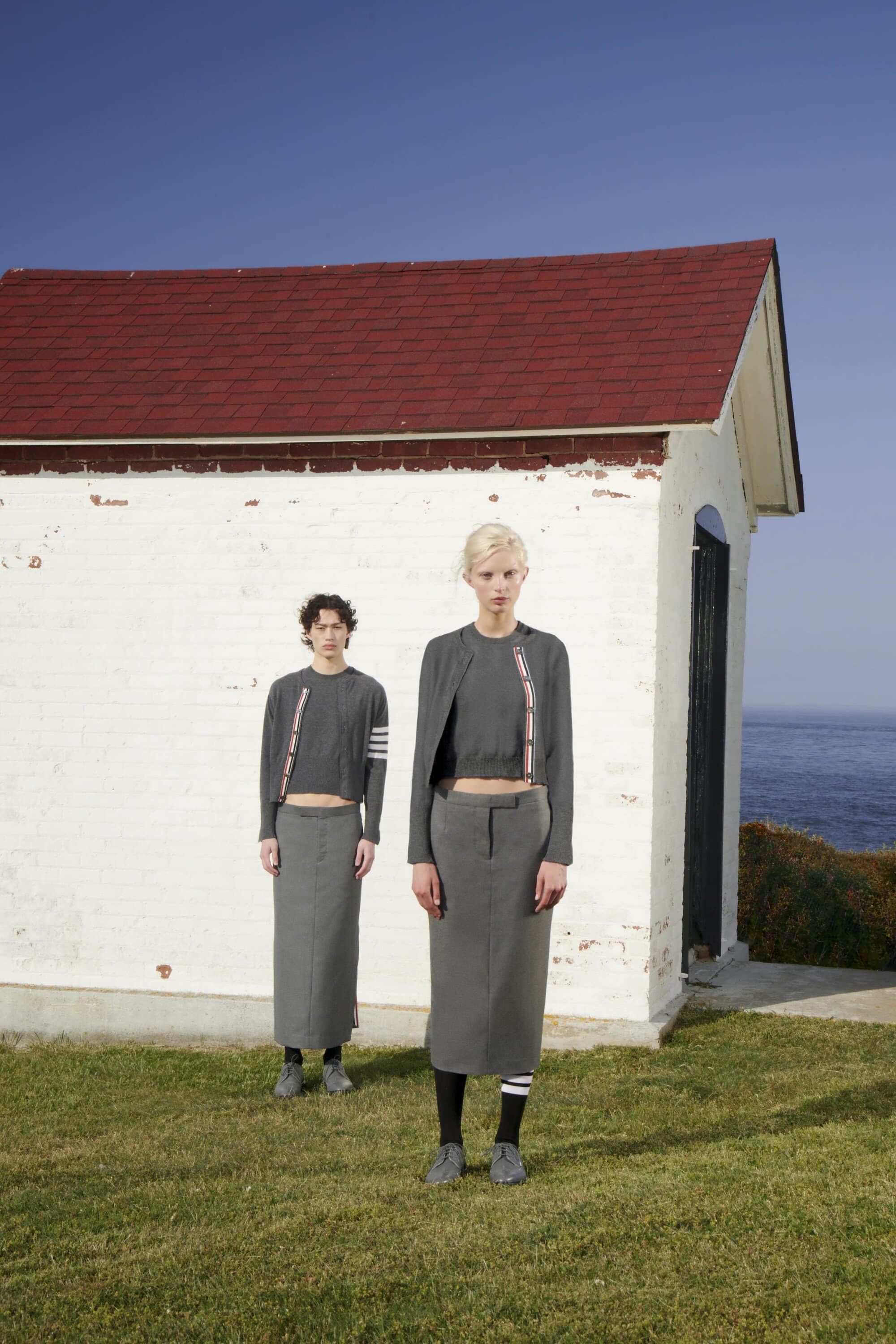 Two fashion models pose in front of a white building near the sea, wearing matching grey tops and skirts with striped accents.