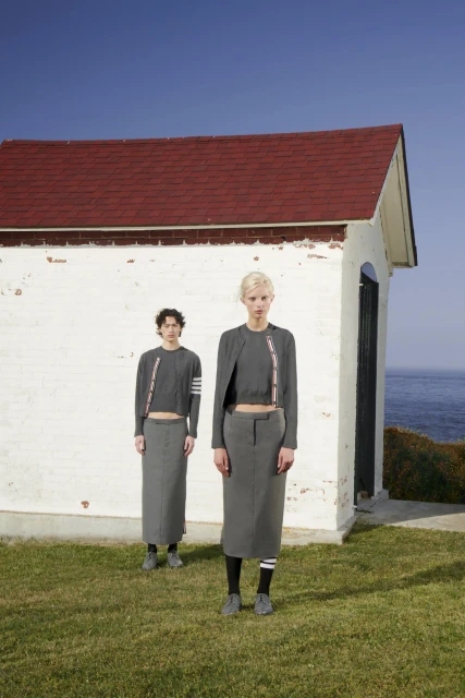 Two fashion models pose in front of a white building near the sea, wearing matching grey tops and skirts with striped accents.