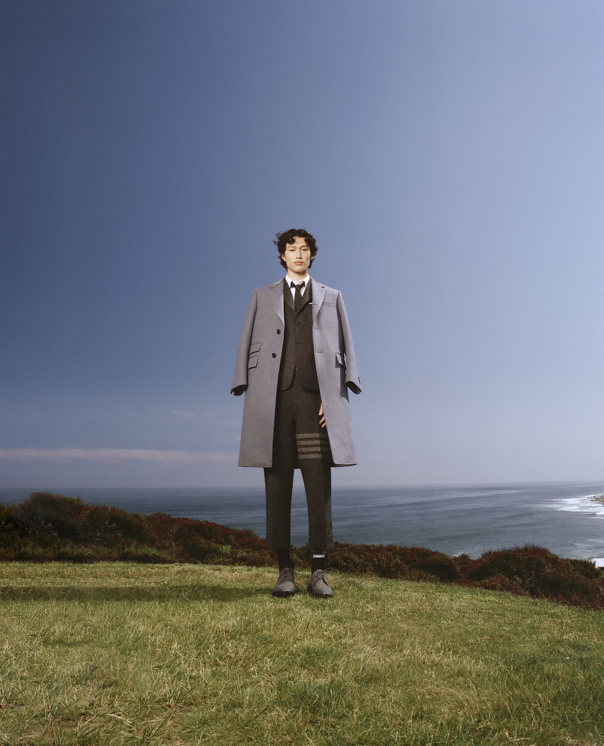 A person stands on a green hilltop overlooking the ocean wearing a suit and overcoat in a fashion or editorial shot.
