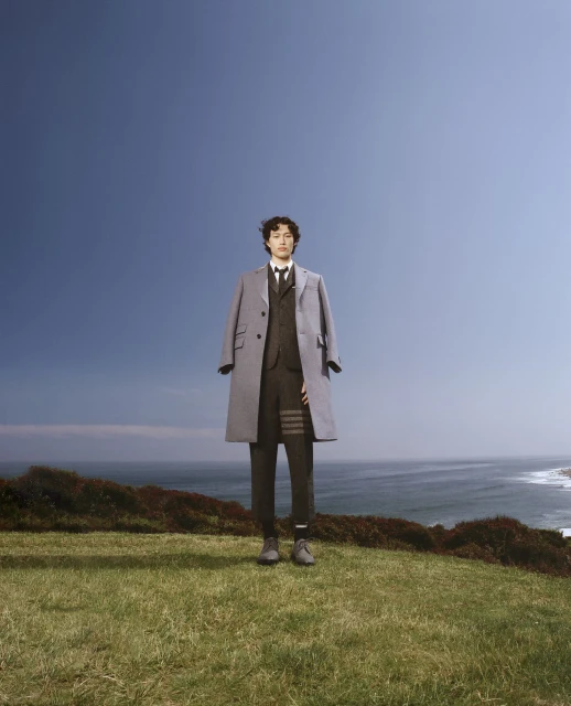 A person stands on a green hilltop overlooking the ocean wearing a suit and overcoat in a fashion or editorial shot.