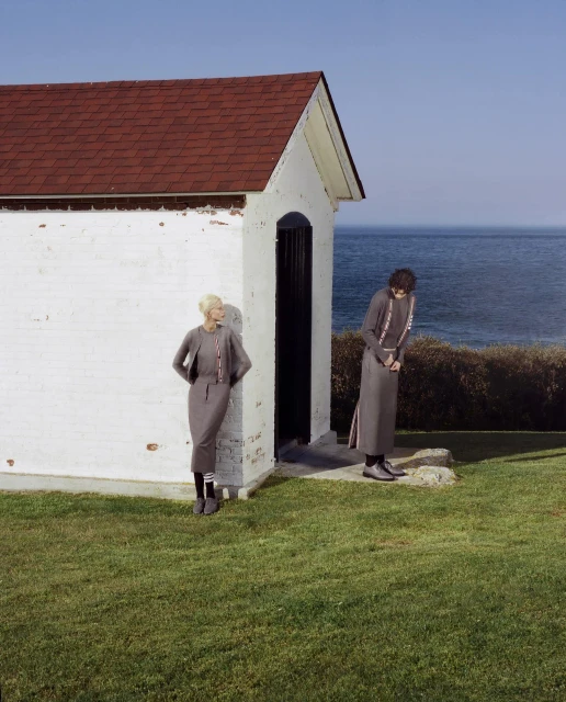 Two women in grey outfits pose near a white building with a red roof by the ocean under a clear sky on a grassy field.