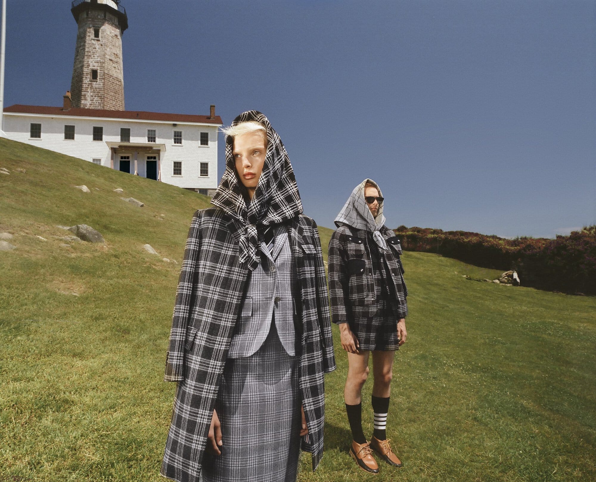 Two women in matching plaid outfits and headscarves stand in a grassy field with a lighthouse and blue sky in the background.