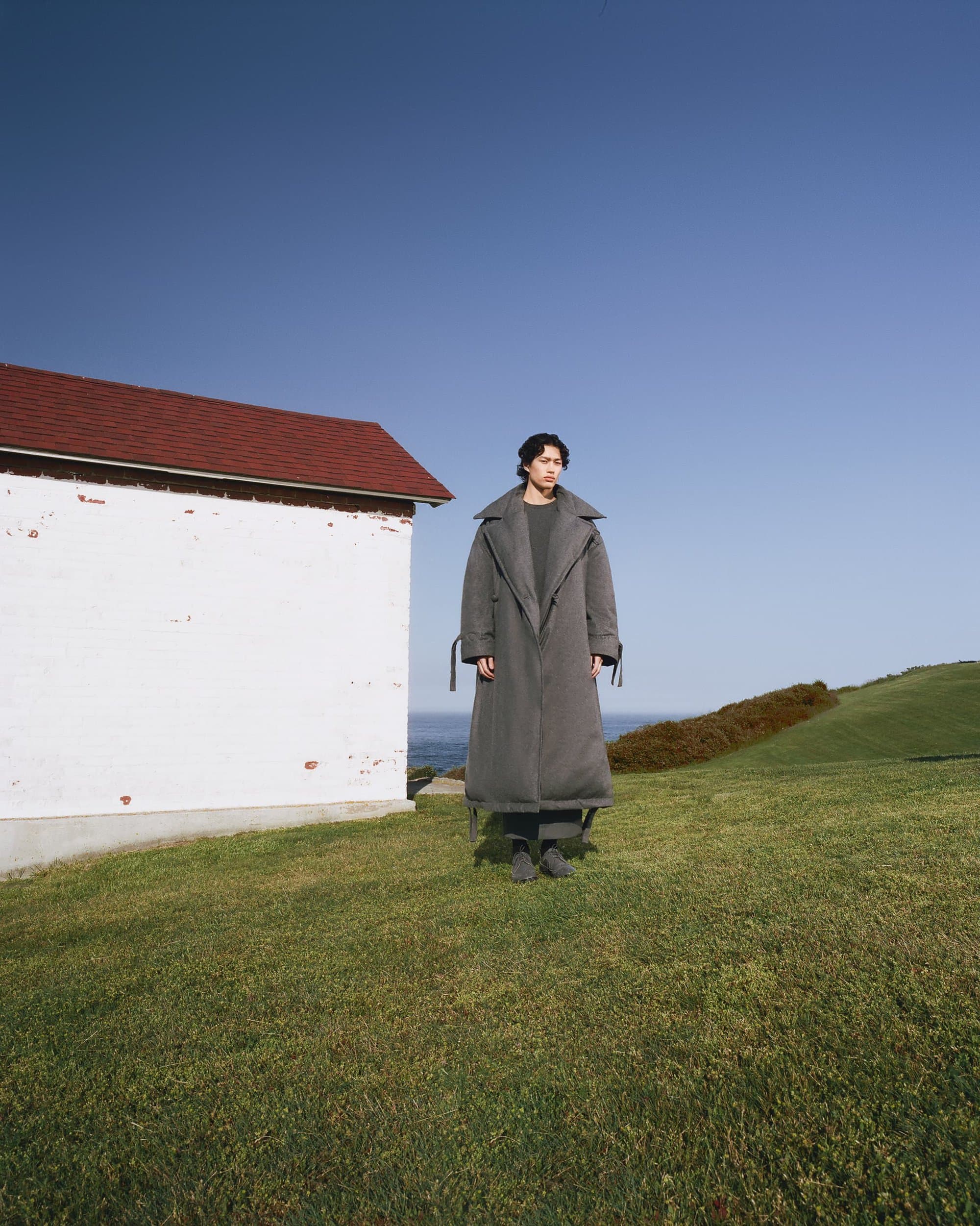 Adult stands in oversized grey coat on green grass near white building with red roof, with clear blue sky and sea in background.