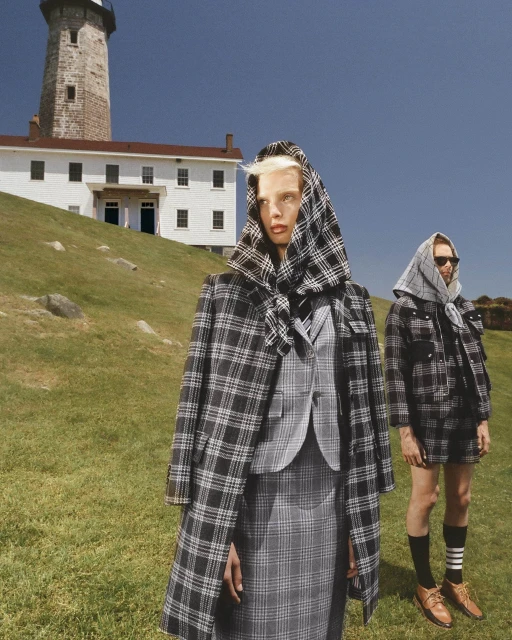 Two fashion models wear matching plaid coats and headscarves near a white lighthouse on a grassy hill under a clear blue sky.