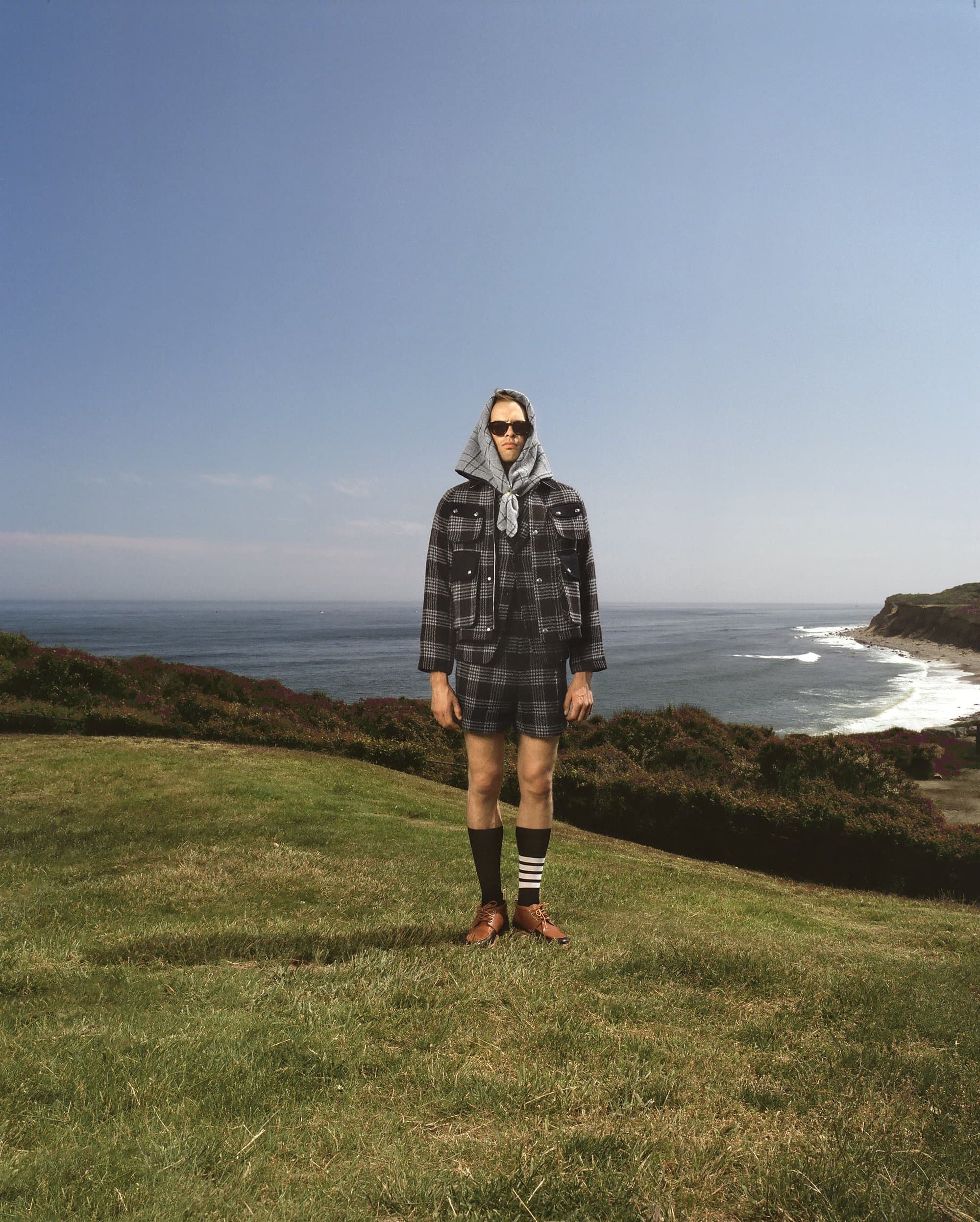 A person stands on a green hill wearing a grey, plaid jacket and shorts, socks with stripes, and brown shoes, with the ocean behind.
