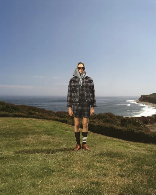 A person stands on a green hill wearing a grey, plaid jacket and shorts, socks with stripes, and brown shoes, with the ocean behind.