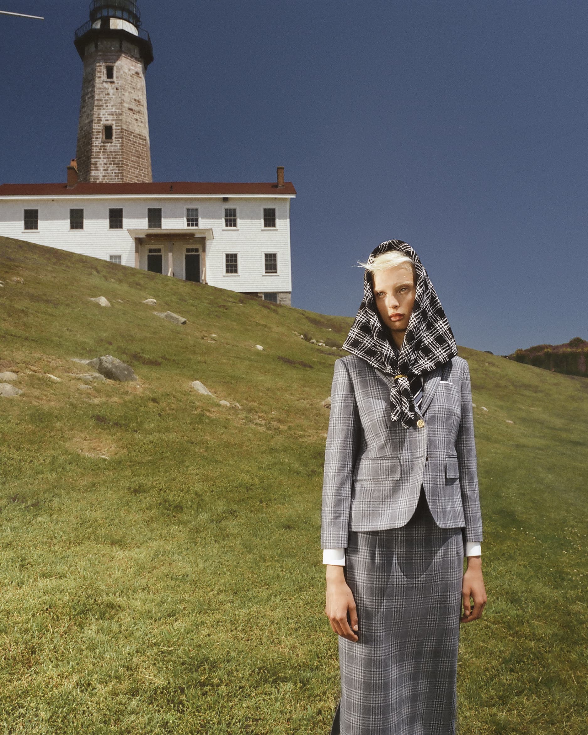 Woman in a grey plaid skirt suit and headscarf stands on a grassy hill with a lighthouse and building in the background on a clear day.