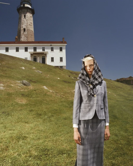 Woman in a grey plaid skirt suit and headscarf stands on a grassy hill with a lighthouse and building in the background on a clear day.