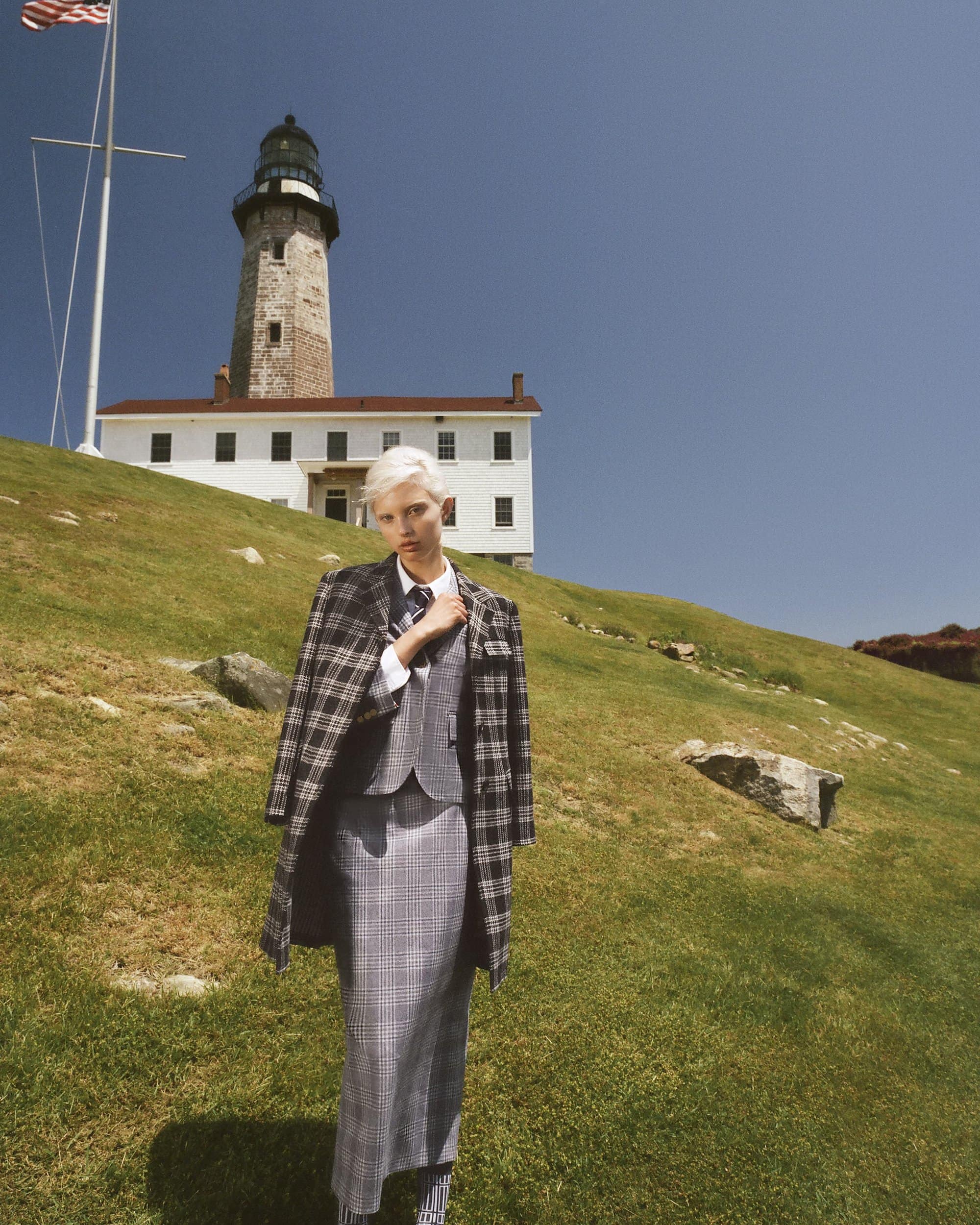 Fashion shot of a woman in a grey plaid skirt suit with a coat slung over her shoulder, standing in a grassy field with a lighthouse.