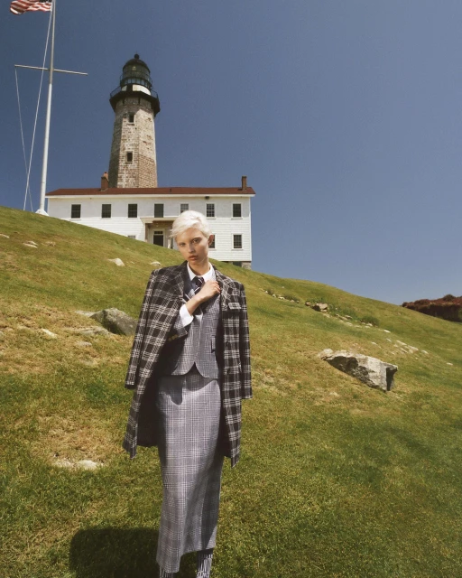 Fashion shot of a woman in a grey plaid skirt suit with a coat slung over her shoulder, standing in a grassy field with a lighthouse.