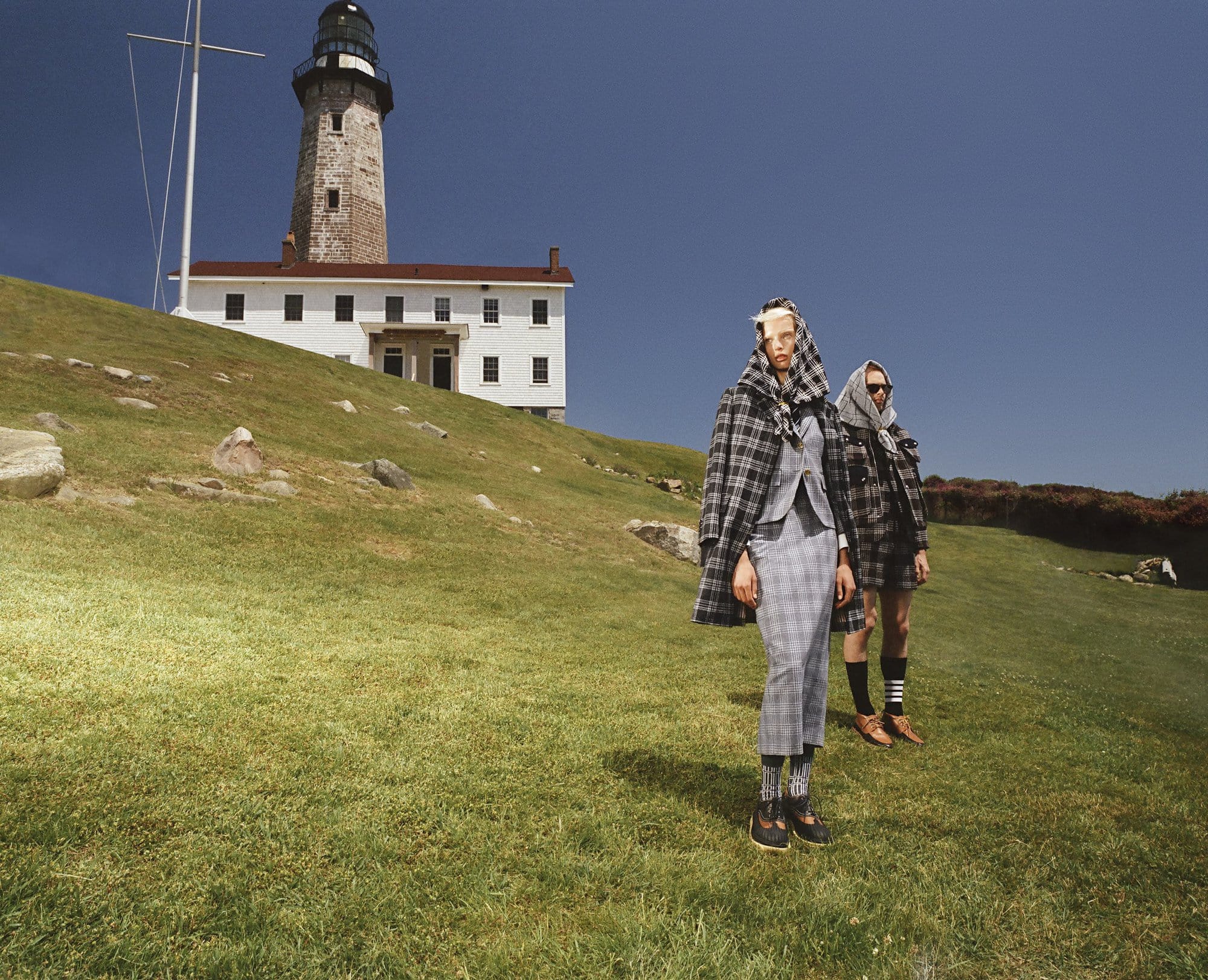 Two models pose on a grassy hill near a lighthouse, wearing grey plaid suits and headscarves under a clear blue sky in daytime.