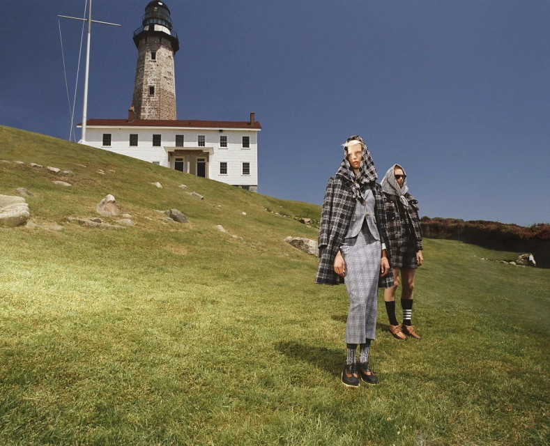 Two models pose on a grassy hill near a lighthouse, wearing grey plaid suits and headscarves under a clear blue sky in daytime.