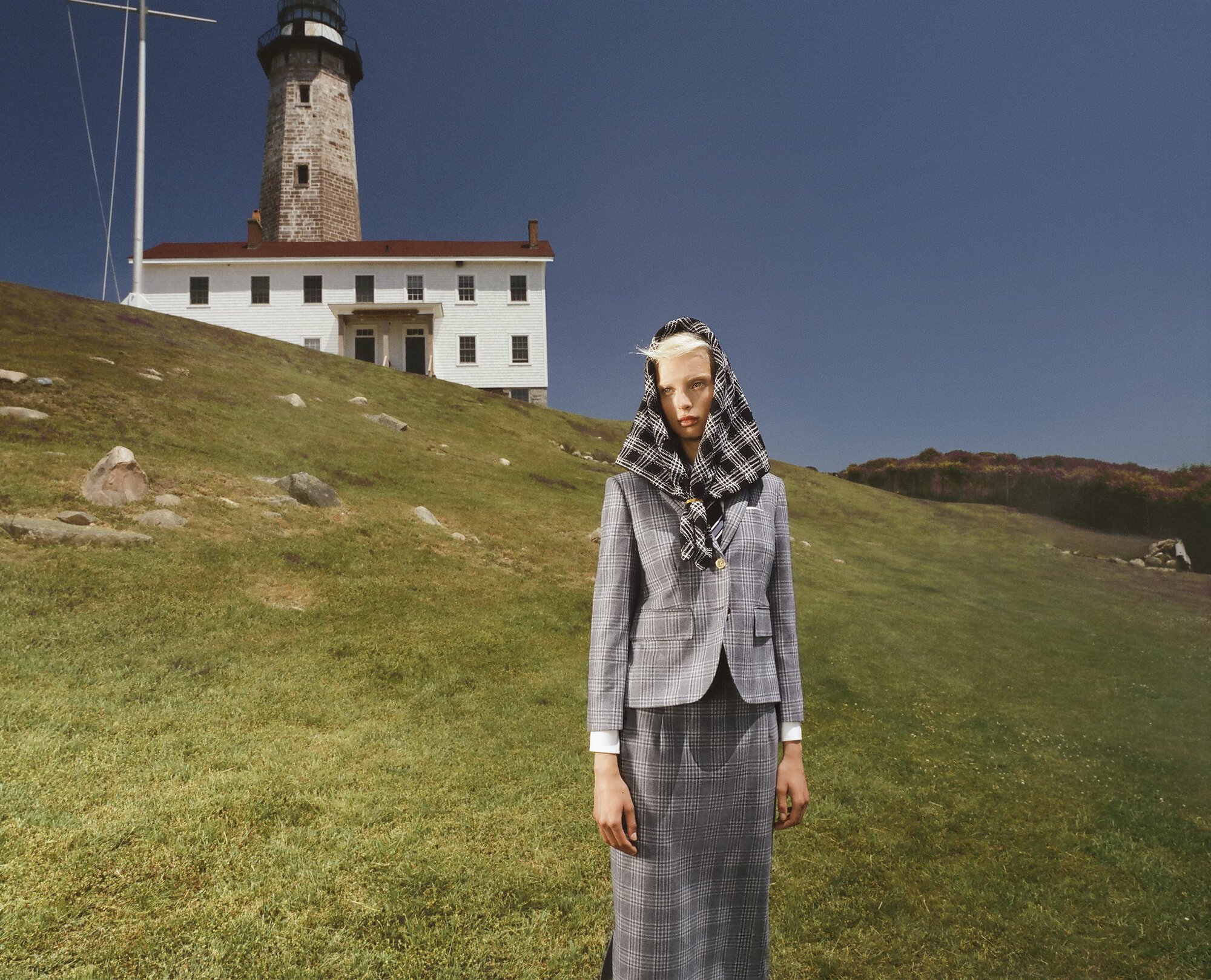 Woman in grey plaid suit with matching headscarf stands in a grassy field with a lighthouse and white building in the background.
