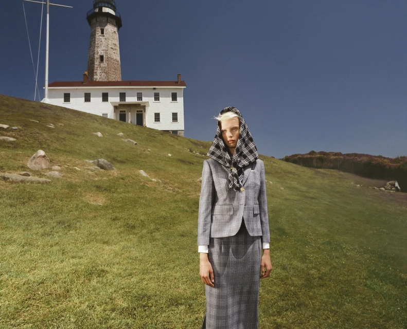 Woman in grey plaid suit with matching headscarf stands in a grassy field with a lighthouse and white building in the background.