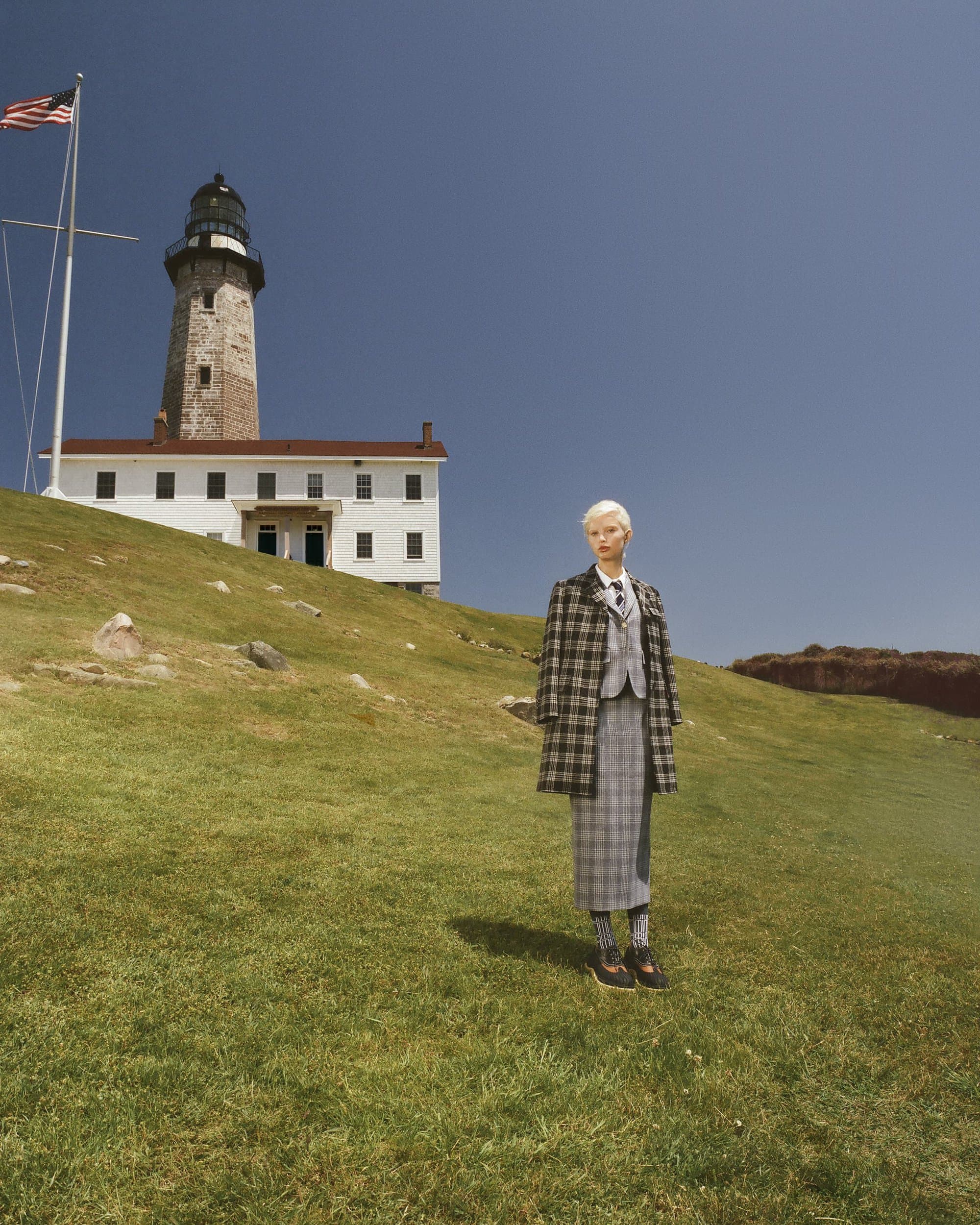 Adult female stands on grassy hillside wearing plaid coat, vest, and skirt, with a lighthouse and American flag visible in the background.