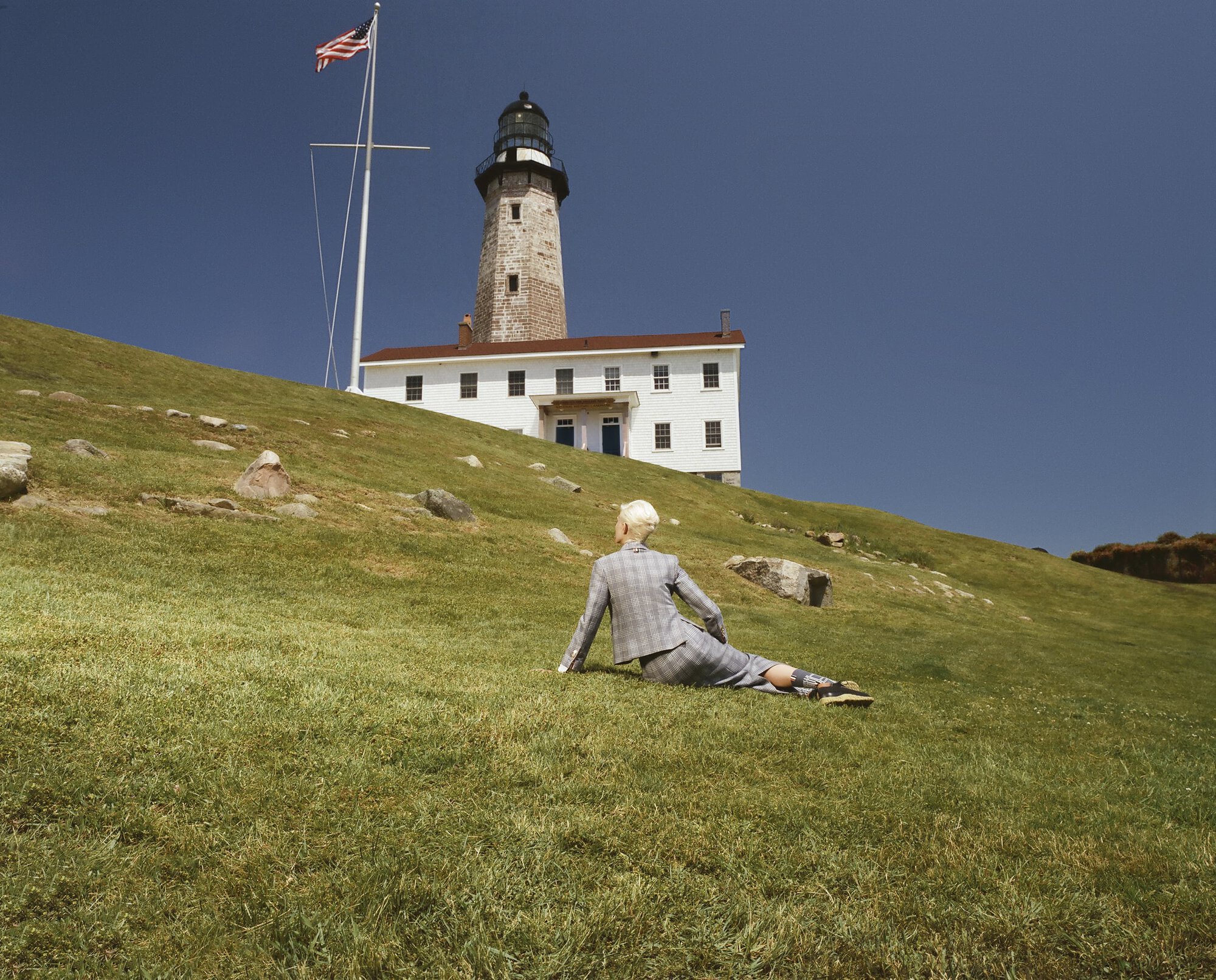 Woman in a grey plaid suit sits on a grassy hill with a lighthouse and the American flag in the background on a sunny day.
