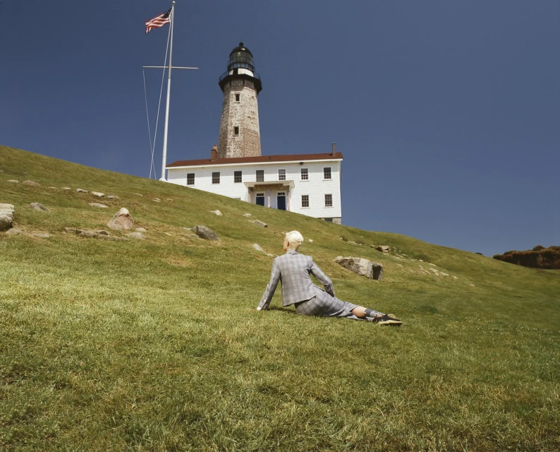 Woman in a grey plaid suit sits on a grassy hill with a lighthouse and the American flag in the background on a sunny day.
