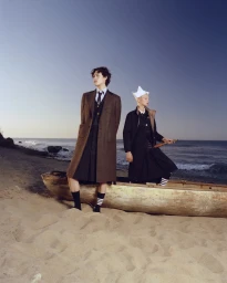 Two models on a sandy beach next to a wooden boat wearing Thom Browne tailored coats and striped socks under a clear sky.